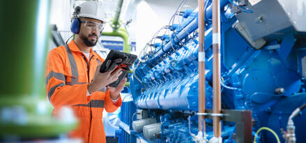 Un ouvrier en combinaison haute visibilité orange et casque inspecte des équipements dans une installation industrielle